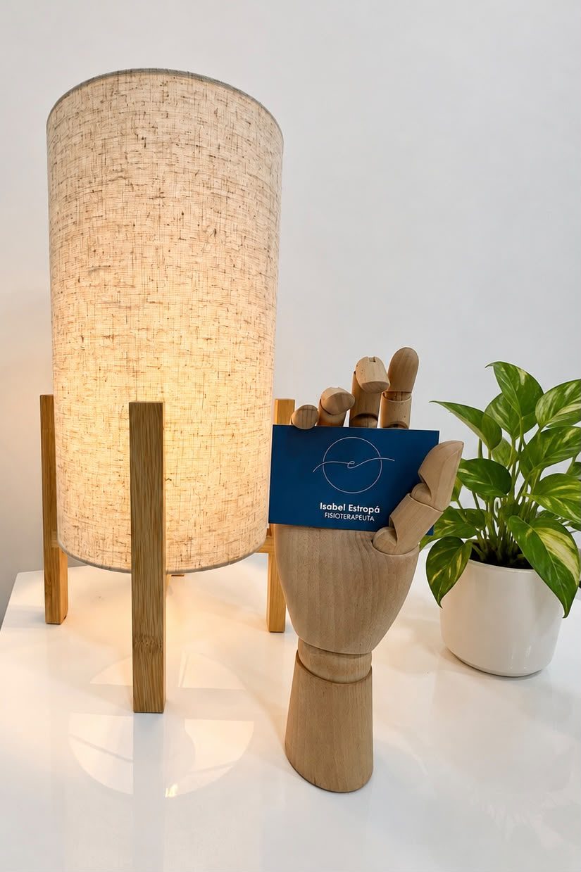 Mano de madera sosteniendo una tarjeta de Isabel Estropá junto a una lámpara y una planta